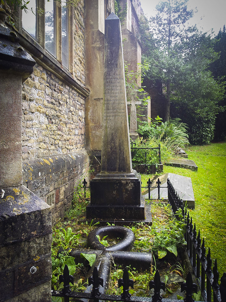 A grave with an obelisk-shaped stone and an Egyptian ankh lying flat in front of it. The grave is flush up against the side of the church