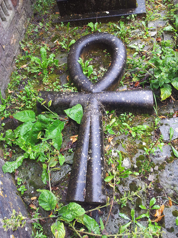 The ankh is made of a dark-coloured stone and is surrounded by greenery which has grown over the grave