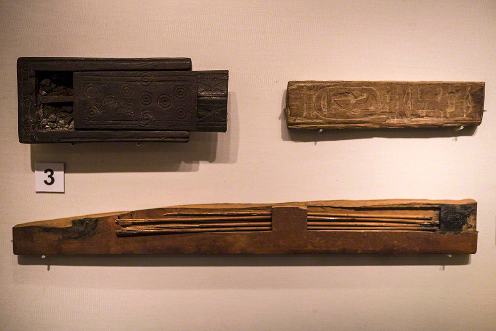 The box is rectangular and black in colour. The lid is partially removed to show the remains of pigment inside. The fragment of palette is rectangular, with hieroglyphs carved into the top. The reed pens are long and thin, four in number, and in a slightly damaged case