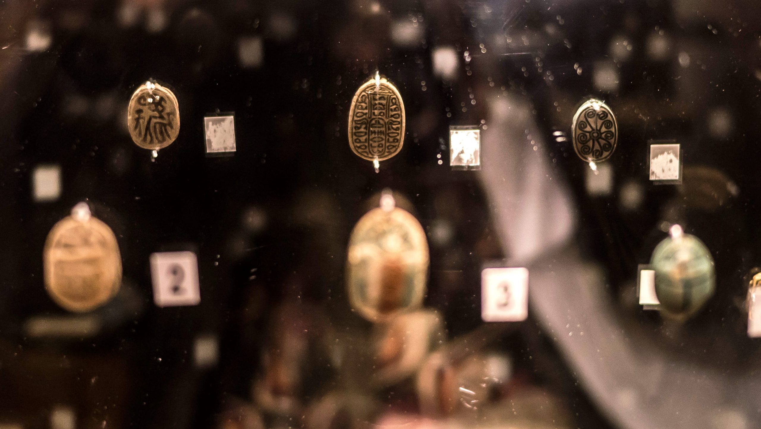 Three small scarab-shaped amulets mounted on a mirror, showing reflections of the backs, which have spiral patterns, stick figures and hieroglyphs