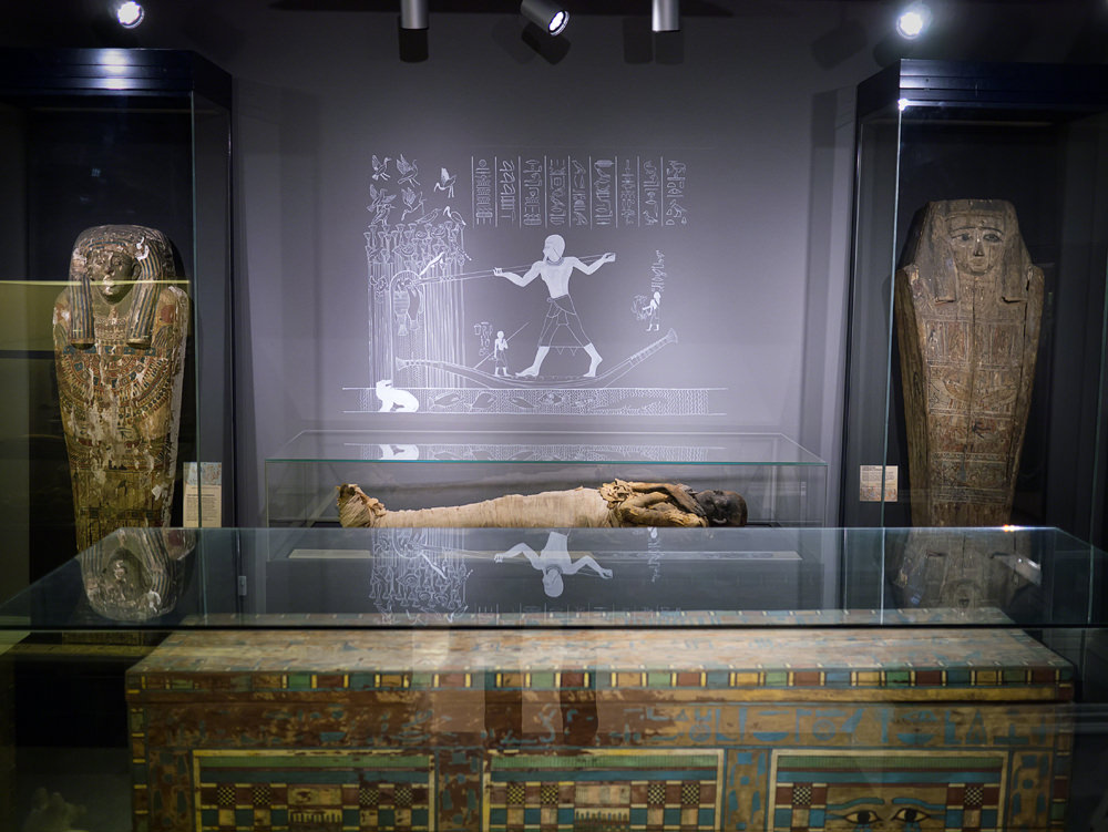 the room has the mummy next to the back wall, with a replica tomb wall relief above him, sarcophagus lids either side and a rectangular, painted coffin in front