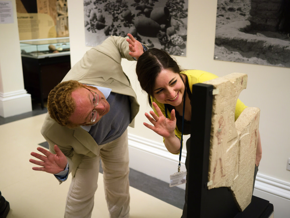 A man and woman bend down to wave comically through a hole in an Egyptian stone inscribed stela