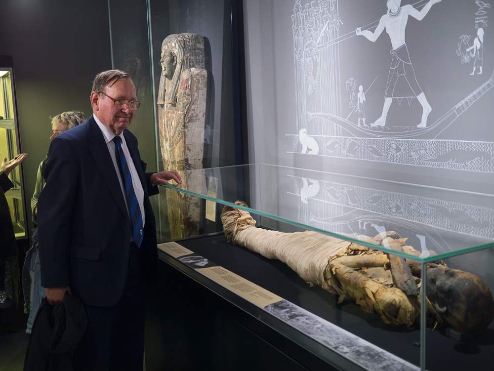 An older man in a smart suit standing and looking at an Egyptian mummy in a glass case