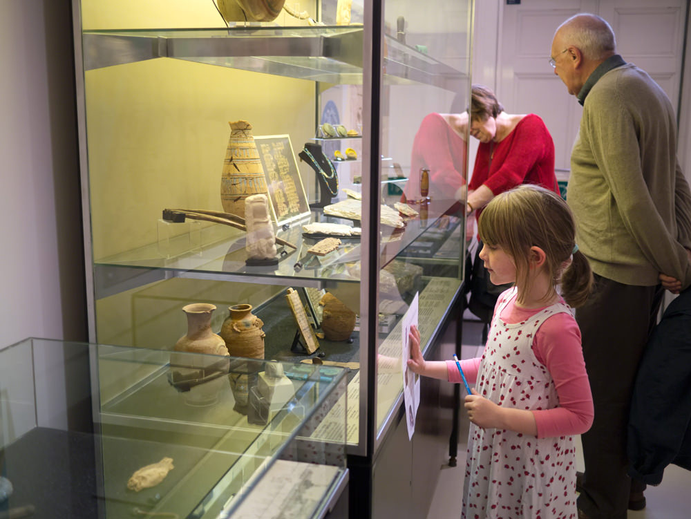 A young girl stands in front of a glass case full of artefacts, with a pen and paper in her hands