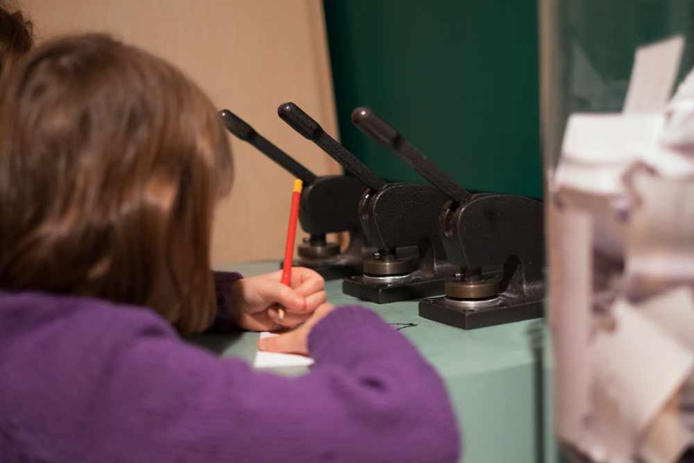 A young girl is writing a message with a pencil. Just in front of her are the three metal embossing stamps. Each one has a lever sticking out the top to work the stamp