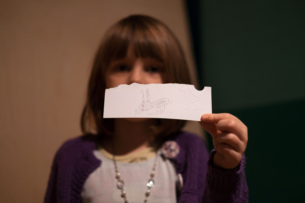 A young girl holds a piece of paper out, partially obscuring her face. She has drawn a rabbit and written 'pet bunny' on it. She has embossed it with a cat