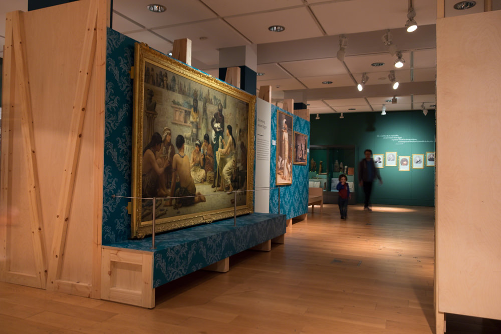 Three large paintings mounted on the wall. The camera is looking at them from an angle, and there are two people walking through the gallery