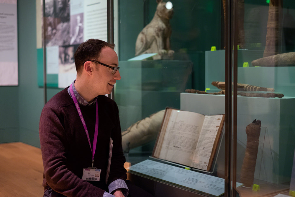 Campbell is kneeling in front of one of the cases, smiling, and looking at the exhibits