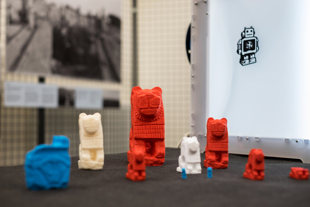 A collection of 3D printed artefacts. There are several lions, ranging in size from around 6 inches tall to less than an inch. There is also a reproduction of a Hathor head. The prints are either red, white or blue in colour.