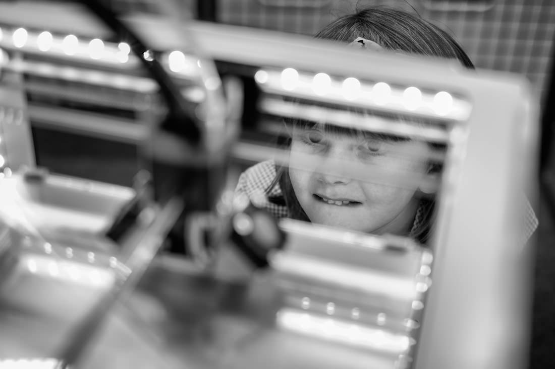 A young girl is standing and watching the 3D printer, with a look of total fascination on her face. The camera is looking down through the front of the printer at her; her head is partially hidden by parts of the printer