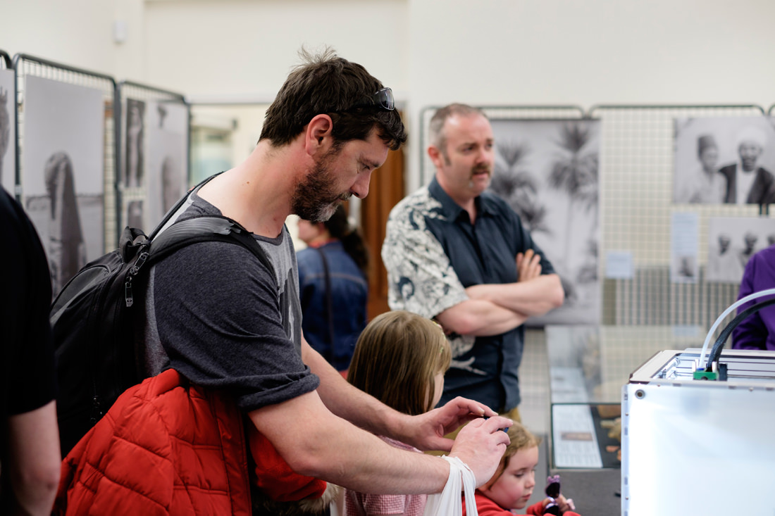 A man is taking a photo of the 3D printer on his phone. There are other people in the room, including two young girls