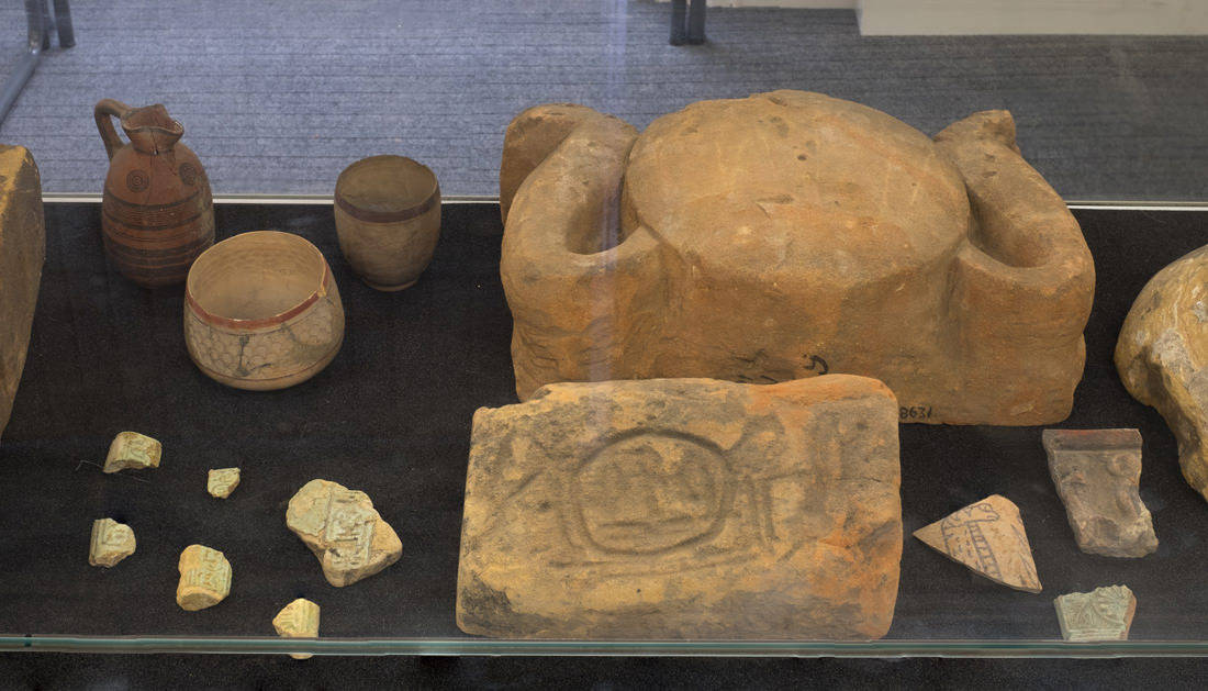 Looking at the artefacts in the centre of the glass display case. There are a couple of larger, stone pieces, one in the shape of a sundisk with cobras on the sides. The other is rectangular with large hieroglyphs inscribed on it. To the left are three fine ceramics and a selection of fragments with hieroglyphs. To the right are a couple more fragments with depictions of cobras.