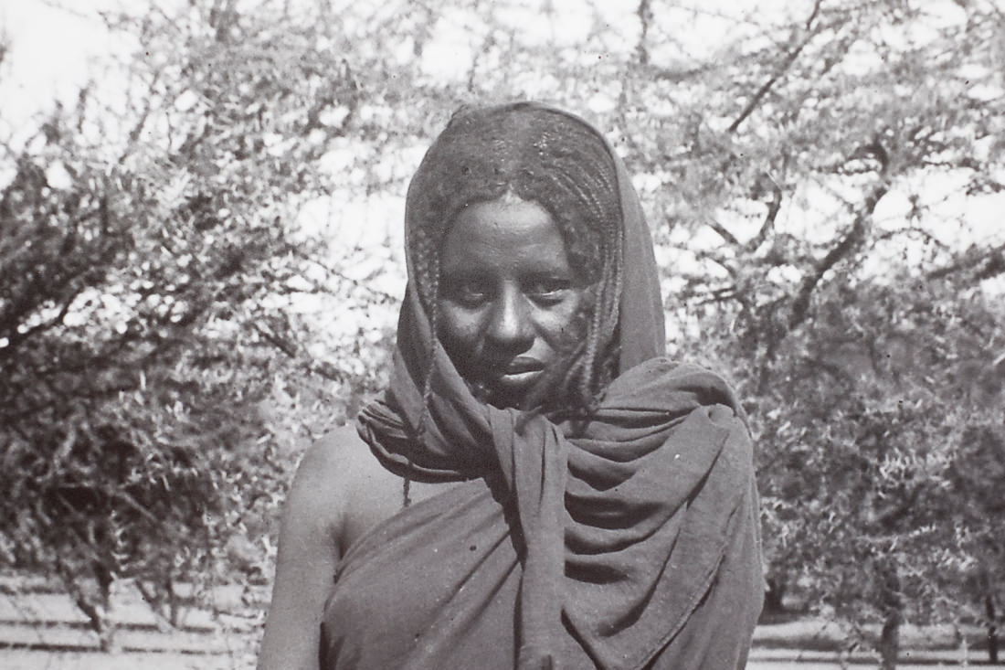 A closeup of one of the photos. It's of a Sudanese woman at the time of the excavations. The closeup crops down to just her head and chest. She's wearing a large piece of cloth draped around her shoulders and head. She's looking directly at the camera. Behind her are trees