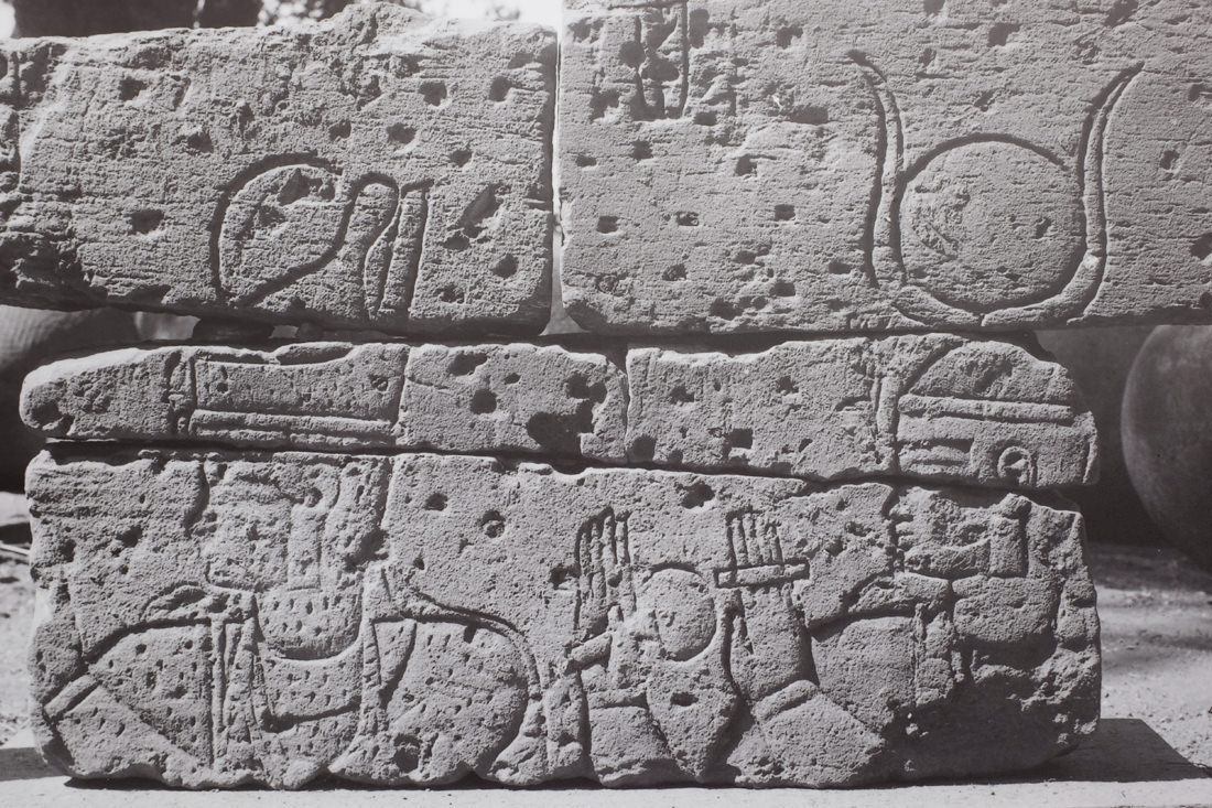 A closeup of a photo of fragments of temple wall pieced back together. It shows the head and shoulders of a king - shown by his wearing the double crown of Egypt - with a goddess behind him. She wears a headdress of a sundisk and cow horns, and she has her hands up in adoration