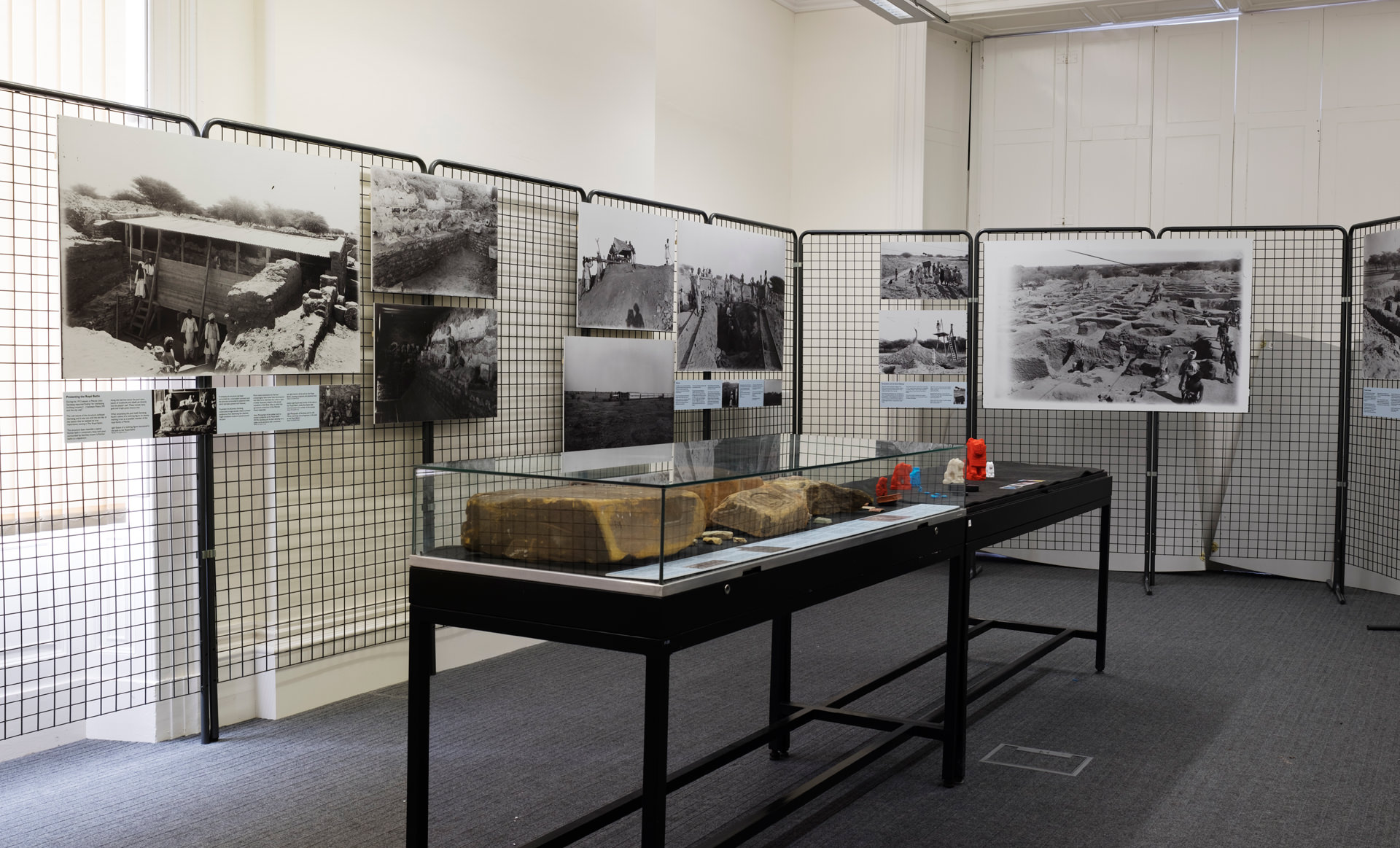 Looking diagonally across the room. The back and left walls are included, which have tall, metal gridded stands to which large reproductions of the excavation photos are attached. In the middle of the room stands a table-sized display case with artefacts inside. Just behind the case is another stand without its glass top with colourful 3D prints of artefacts on top