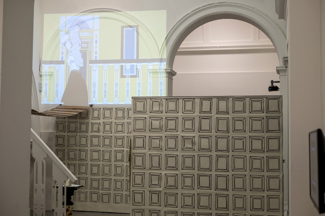 A hallway with mock wooden panelling made of paper with squares drawn along them. At the top left is a cartoon projected onto the wall of a Victorian-era maid carrying piles of laundry