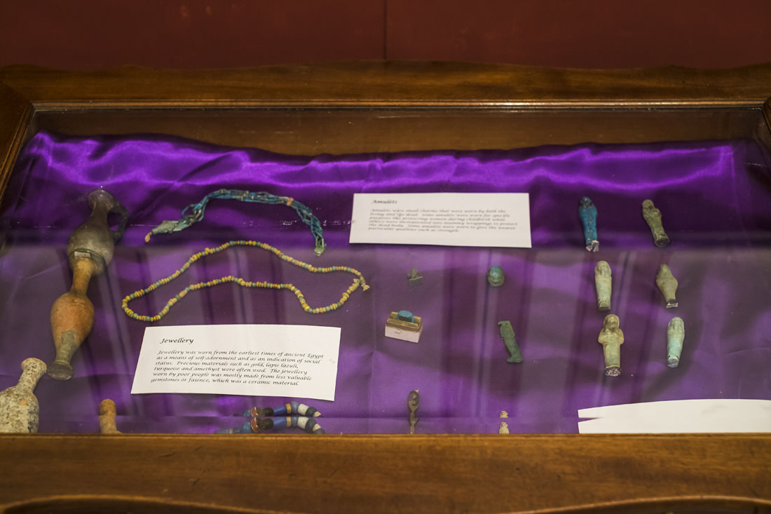 A wooden display case, to be viewed from the top. It's lined with a shiny purple fabric on which lies pieces of Egyptian jewellry and other small pieces, such as shabtis and amulets