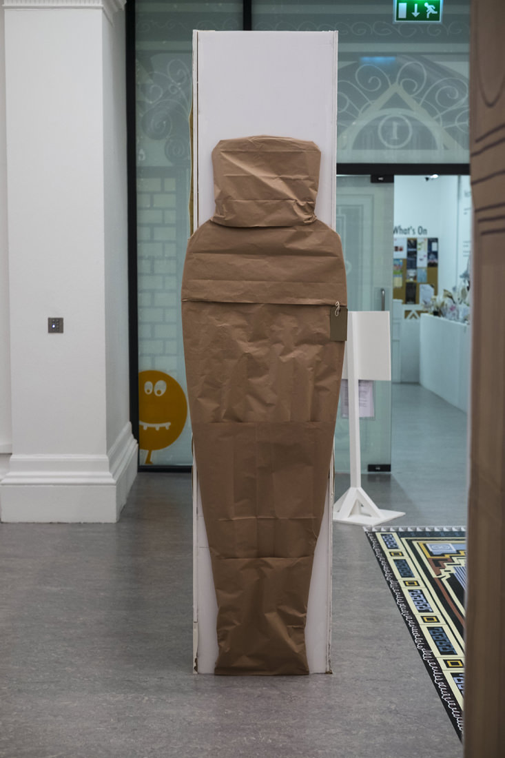 An anthropoid sarcophagus made of brown paper standing against the end of a wall