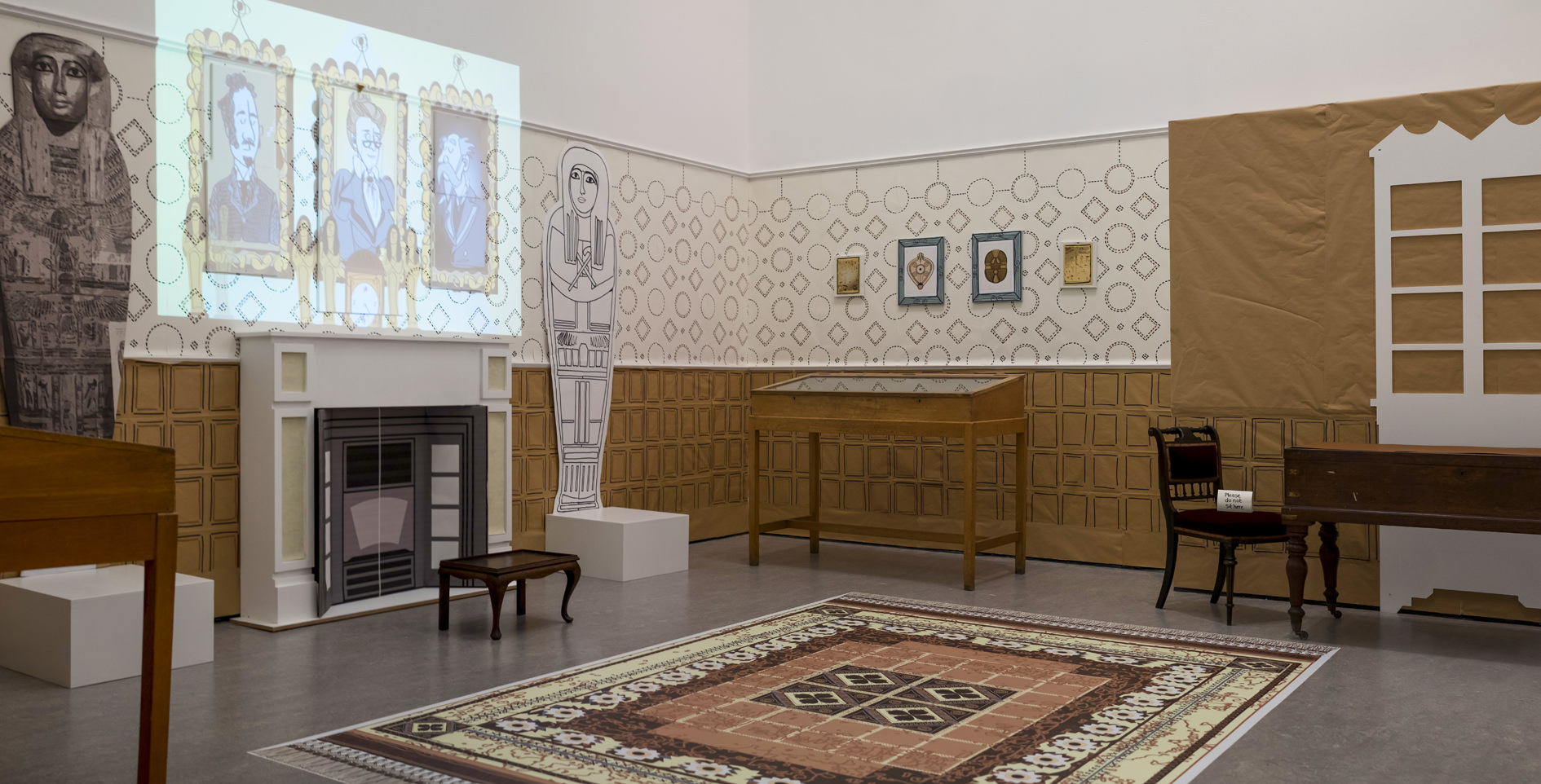 A large room with a fireplace, table and chair and a couple of wooden display cases containing artefacts. There are paper mockups of Egyptian coffins either side of the fireplace and cartoon-style portraits being projected onto the wall above the fireplace