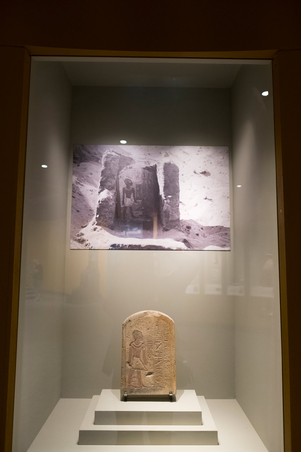 A carved, painted funerary stela in a display case with an old photo of it in situ when it was first discovered above it