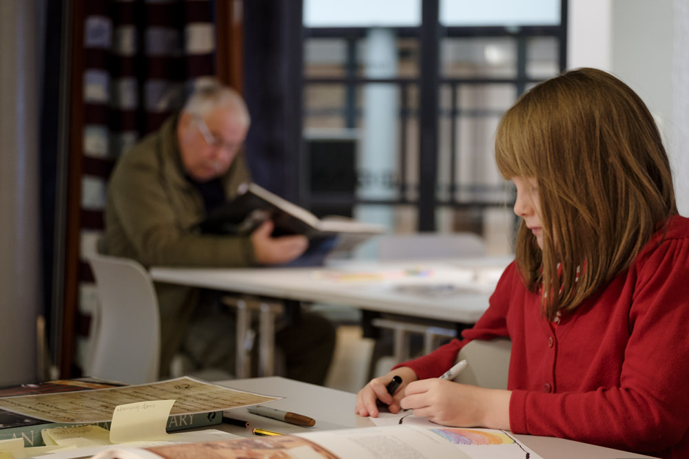 A girl sitting and drawing, with an elderly man in the background, reading a book