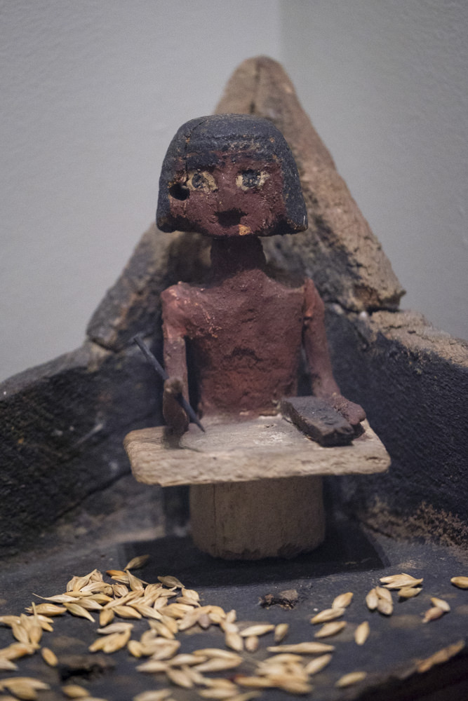 An ancient Egyptian wooden scribe sitting in the corner of a granary with his pen, palette and paper on his knees