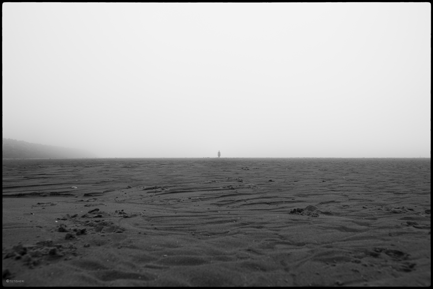 an expanse of textured sand on a foggy day with an anthony gormley statue in the distance