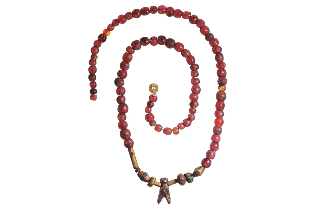 The necklace is arranged in a spiral. It is mostly small, round carnelian beads with a small fly pendant in the middle, flanked by a few beads of assorted shapes and materials.