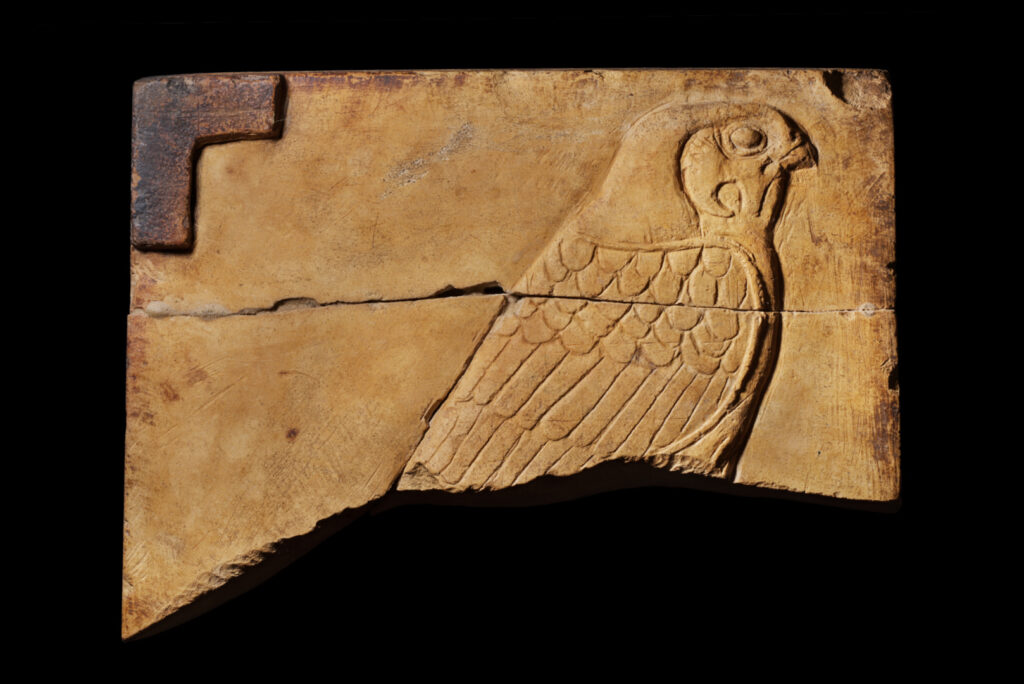 A broken rectangle of yellow stone with the top half of a carved falcon. The bird has detail carved into the face and wing.