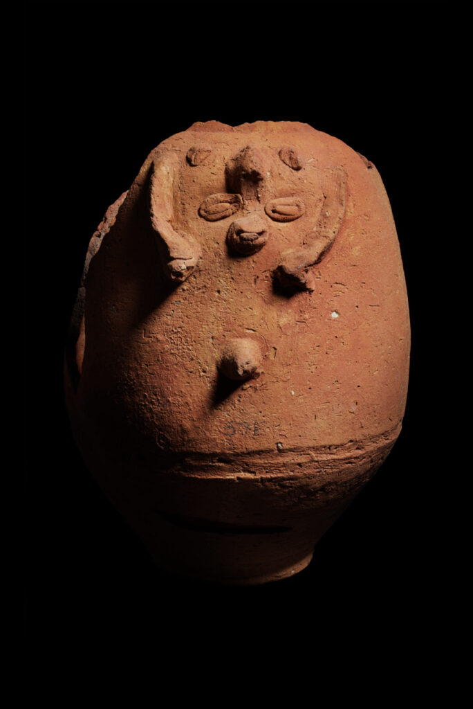 A rough, ceramic pot of the Egyptian god Bes. His facial features are made from hand-sculpted pieces adhered to the pot.