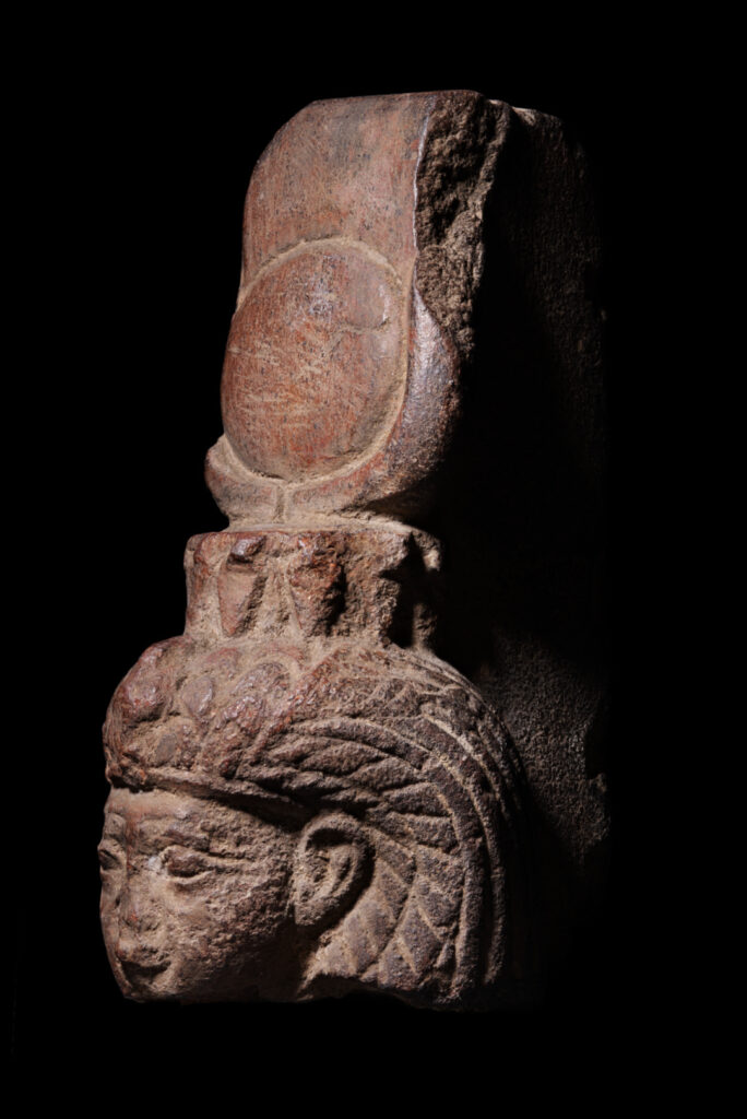 The head from a figurine of an Egyptian goddess. She has a tall headdress with a sundisk and crescent moon.