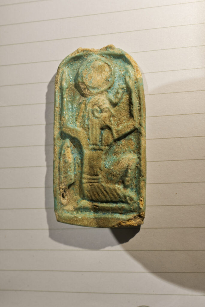 A round-topped rectangular faience amulet with a depiction of the god Shu, kneeling with his hands up. He has a sundisk headdress and a feather by his side.