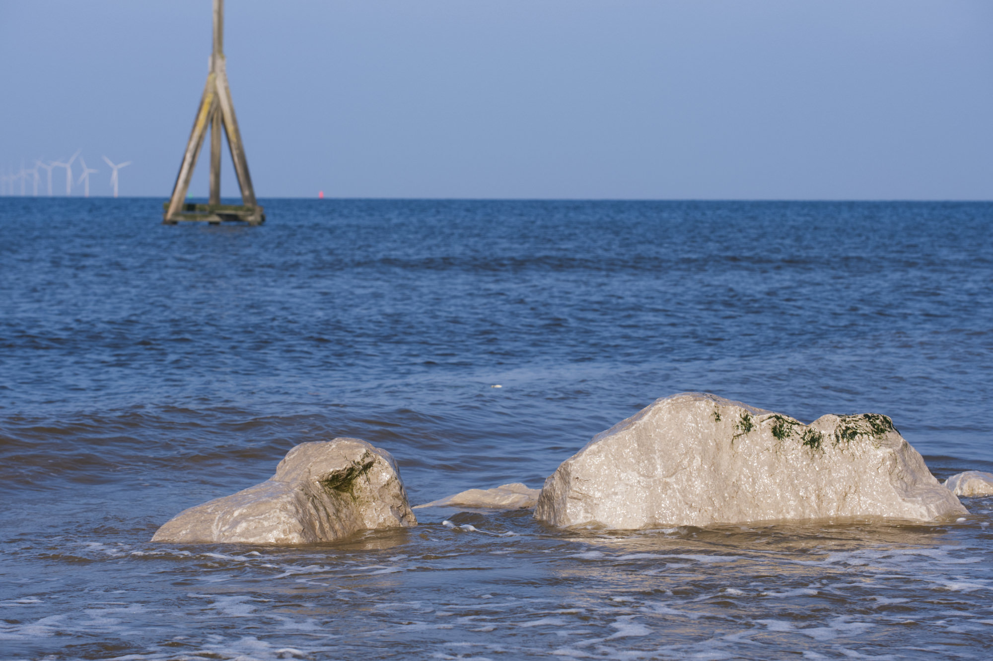 Some small, grey rocks in the sea. In the background is a tall wooden pole structure and some wind turbines in the far distance.