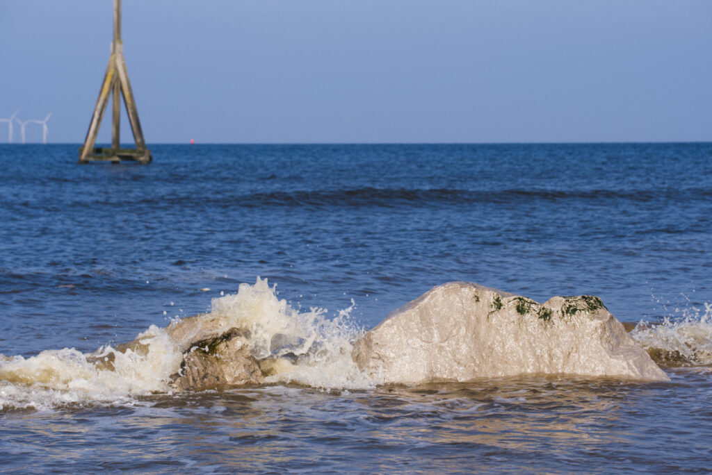 Some small, grey rocks in the sea with a small wave breaking over them. In the background is a tall wooden pole structure and some wind turbines in the far distance.
