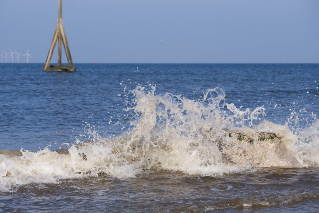 Some small, grey rocks in the sea with a medium wave breaking over them. In the background is a tall wooden pole structure and some wind turbines in the far distance.