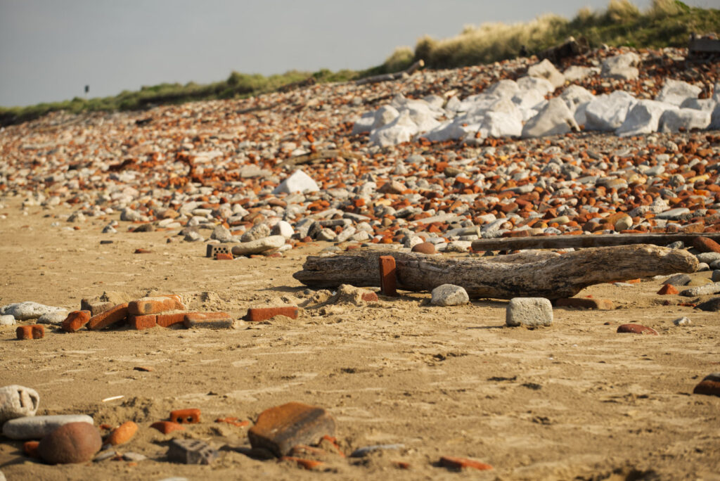 The beach with sand in the foreground and piles of bricks, rocks and stones heading off into the distance. In the middle is a large piece of driftwood.