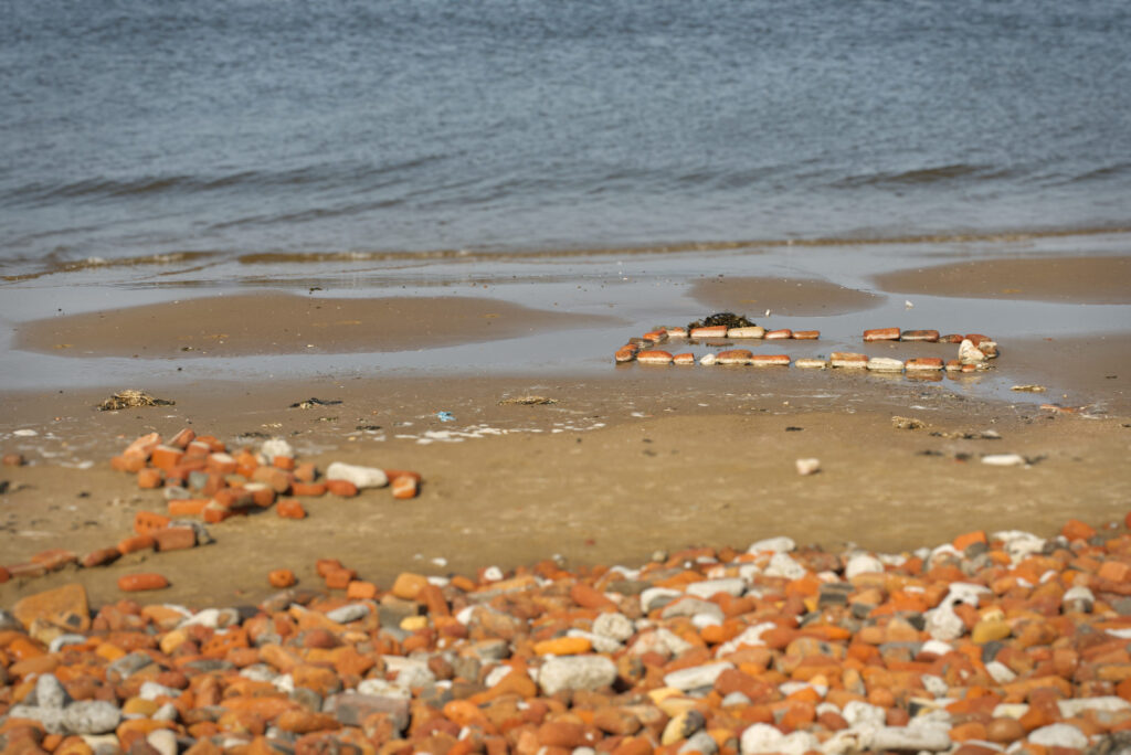 A view of the beach looking from piles of bricks, across some sand and down to the water. On the sand, just by the water's edge, is a rectangle made from a single layer of old bricks. There's a gap in the bricks on one of the long sides of the rectangle.